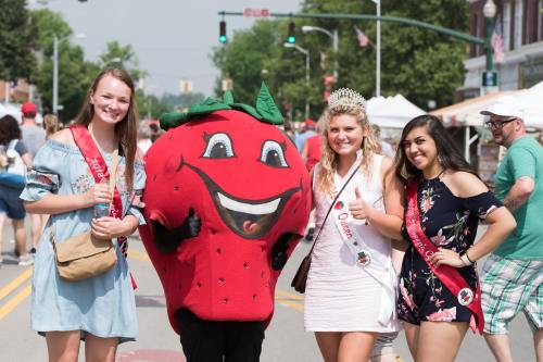 Three young women in festive attire pose with a large strawberry mascot at a lively outdoor event.