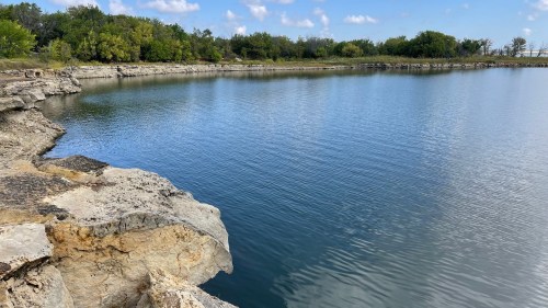 A calm body of water surrounded by rocky shores and greenery under a blue sky with scattered clouds.