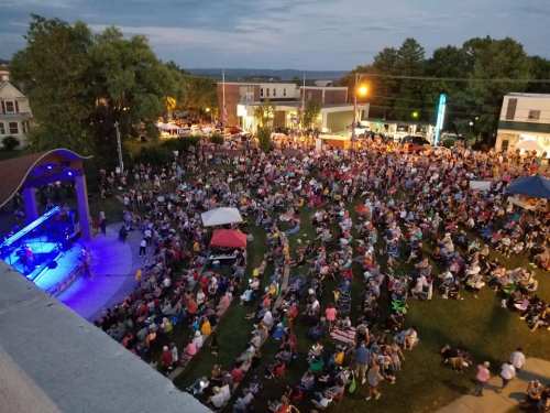 A large crowd gathers in a park for an outdoor concert as dusk settles, with stage lights illuminating the scene.