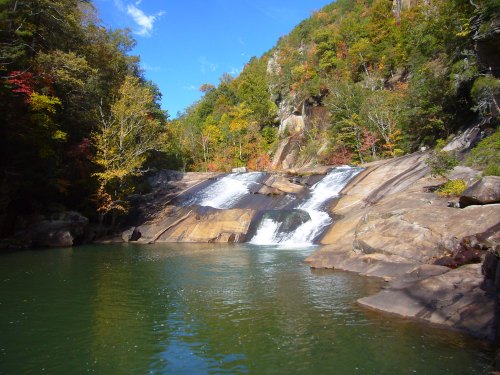 A serene waterfall cascades over smooth rocks into a clear pool, surrounded by vibrant autumn foliage and blue skies.