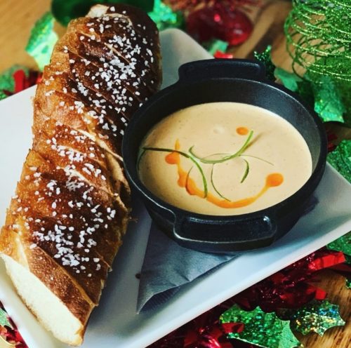 A pretzel with coarse salt served alongside a small bowl of creamy dipping sauce, garnished with green onions and drizzle.