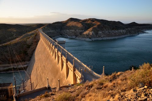 A dam stretches across a reservoir, surrounded by hills and a clear sky during sunset.