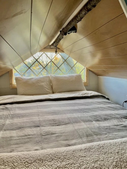Cozy loft bedroom with a sloped ceiling, large window, and a neatly made bed with white pillows and a striped blanket.