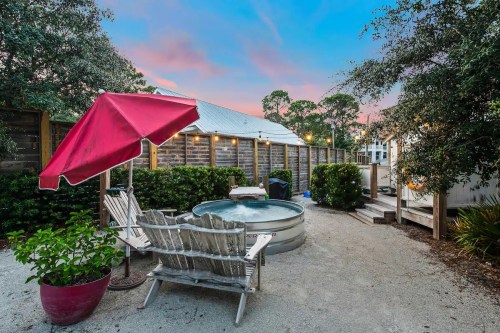 A cozy outdoor space featuring a hot tub, wooden chairs, a red umbrella, and lush greenery under a colorful sky.