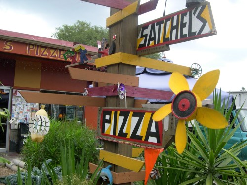 Colorful wooden signpost with directional arrows for "Satchel's" and "Pizza," surrounded by greenery and playful decorations.