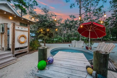 A cozy outdoor space with a hot tub, wooden seating, colorful floats, and string lights against a sunset sky.