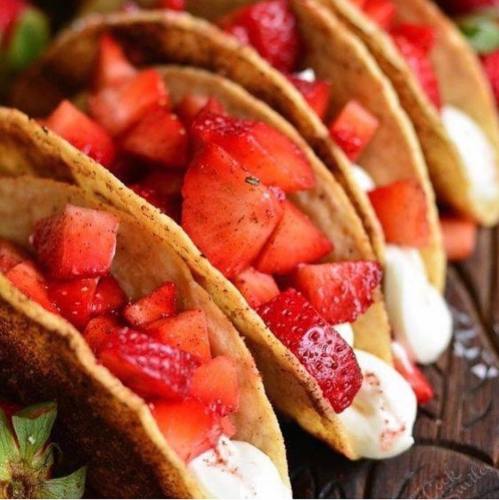 Four dessert tacos filled with fresh strawberries and cream, arranged on a wooden surface.