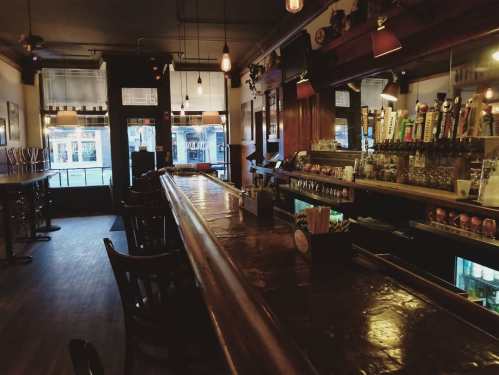 A dimly lit bar with a polished wooden counter, beer taps, and empty stools, creating a cozy atmosphere.
