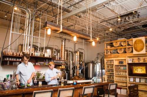 Two bartenders prepare drinks at a stylish distillery bar with metal equipment and wooden shelves in a bright, industrial space.