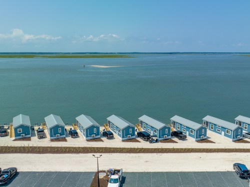 Aerial view of colorful waterfront cabins lined along a beach, with parked cars and a calm waterway in the background.
