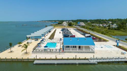 Aerial view of a waterfront area with a pool, parking, and beach houses along the shore under a clear blue sky.