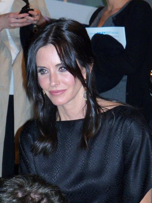 A woman with long dark hair and earrings smiles while seated, surrounded by people in a social setting.