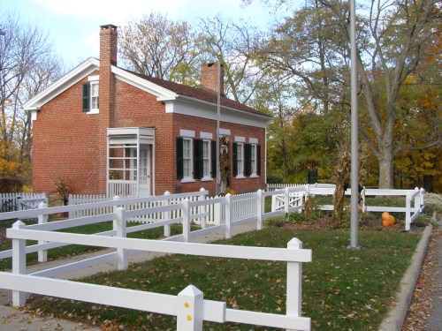 A brick house with white trim, surrounded by a white picket fence and autumn foliage, featuring pumpkins in the yard.