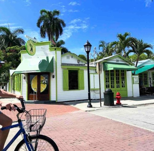 A cyclist rides past a colorful, tropical-themed building with green accents and palm trees under a clear blue sky.