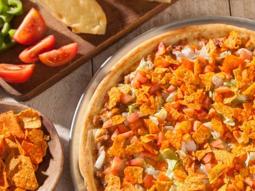 A close-up of a pizza topped with lettuce, tomatoes, and crunchy chips, alongside sliced vegetables on wooden platters.