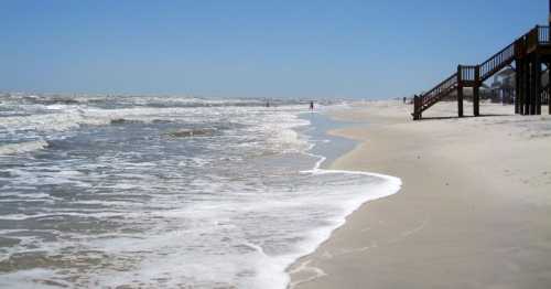 A sandy beach with gentle waves, a clear blue sky, and a wooden staircase leading to a beach house in the distance.