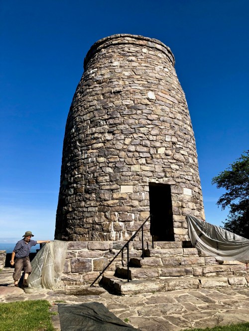 A stone tower stands against a clear blue sky, with a person preparing a tarp nearby.