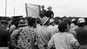 A crowd of people gathered outdoors, with two individuals speaking and holding a large map or poster.