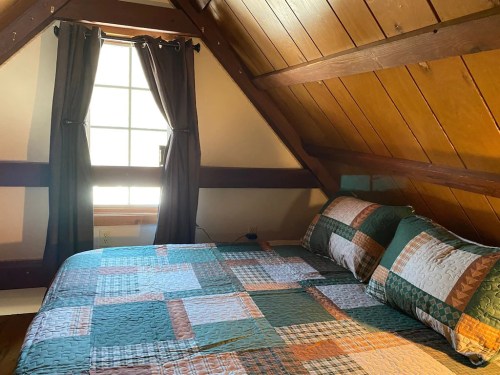 Cozy attic bedroom with a quilted bedspread, wooden walls, and a window with dark curtains.
