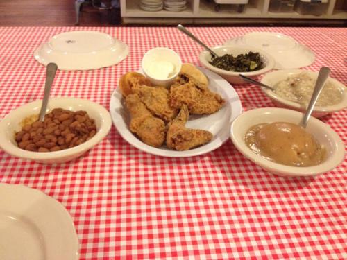 A table set with fried chicken, sides of beans, greens, and a dessert, all on a red and white checkered tablecloth.