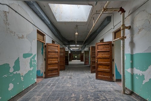Abandoned hallway with peeling paint, open wooden doors, and a skylight illuminating the space.