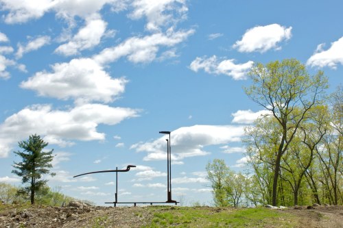 A large metal sculpture stands against a blue sky with fluffy clouds, surrounded by trees and rocky terrain.