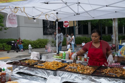 A vendor in a red apron cooks food at a bustling outdoor market, with people and greenery in the background.