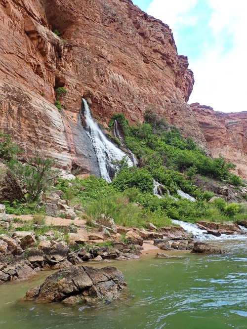 A waterfall cascading down a rocky cliff into a green river, surrounded by lush vegetation and red rock formations.