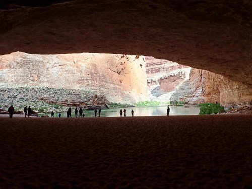 Silhouetted figures stand at the entrance of a large cave, overlooking a river surrounded by rocky cliffs.