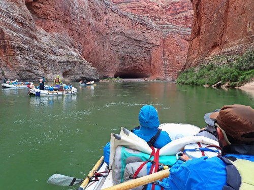 A group of people in rafts navigating a river surrounded by towering red rock cliffs and lush greenery.