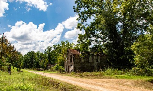 A dirt road leads to an old, abandoned house surrounded by trees and a bright blue sky with fluffy clouds.