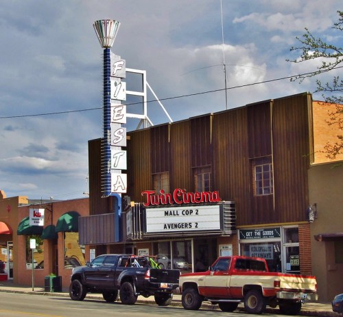 A vintage cinema building with a marquee displaying "Mall Cop 2" and "Avengers 2," surrounded by parked trucks.