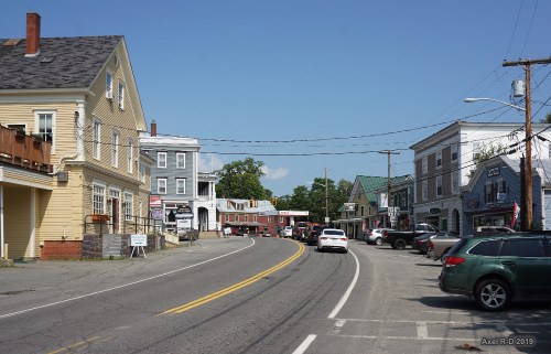 A small town street lined with buildings, shops, and parked cars under a clear blue sky.