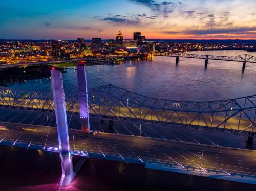 Aerial view of a bridge over a river at sunset, with city lights reflecting on the water and a colorful sky.