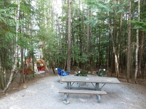 A campsite in a forest with a picnic table, camping chairs, and a tent among tall trees.