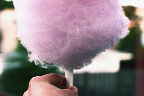 A hand holding a fluffy pink cotton candy on a stick, with a blurred background.