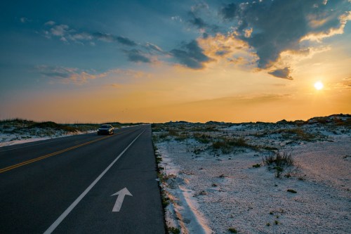 A car drives along a road beside sandy dunes under a colorful sunset with scattered clouds.