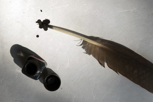 A quill feather beside a small ink bottle and a pile of ink powder on a textured surface.