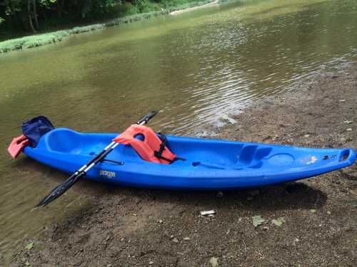 A blue kayak with a red life jacket rests on the shore of a calm river, surrounded by greenery.