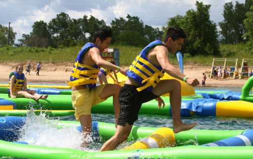 Two people in life vests jump on inflatable water obstacles at a beach, splashing water around them.