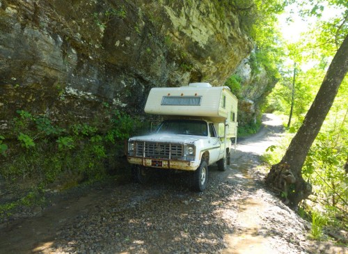 An old white camper parked beside a rocky cliff on a narrow, tree-lined dirt road.