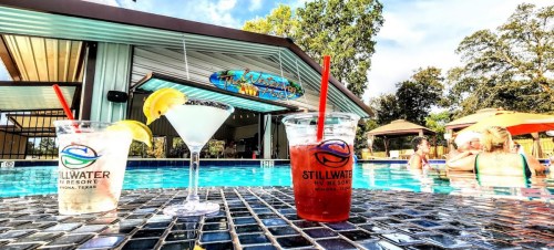 Two colorful cocktails sit on a table by a pool, with a resort bar and people enjoying the sun in the background.
