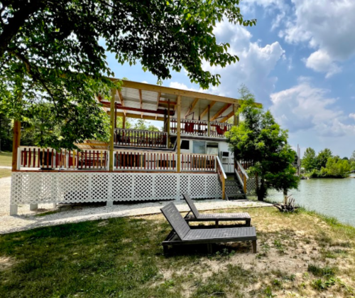 A lakeside cabin with a spacious deck, surrounded by trees and two lounge chairs on the grassy shore.