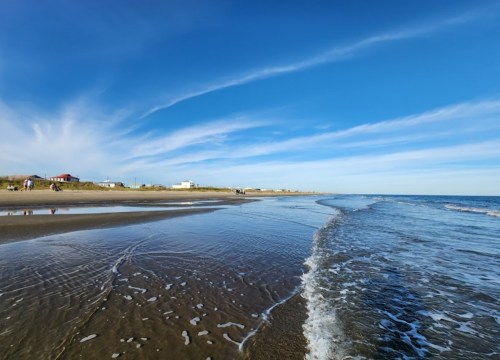 A serene beach scene with gentle waves, a clear blue sky, and distant buildings along the shoreline.