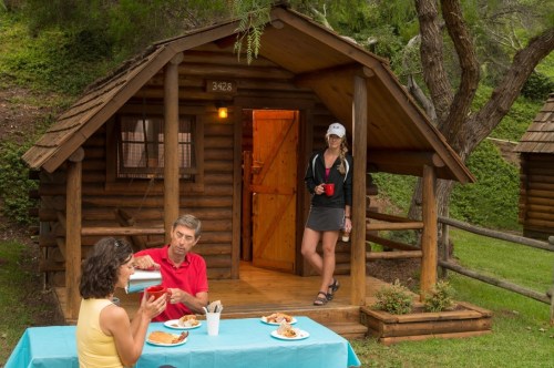 A couple enjoys a meal outside a log cabin while a woman stands at the entrance, holding a drink.