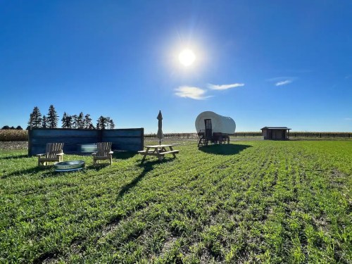 A sunny farm scene with a covered wagon, picnic table, and chairs on green grass, surrounded by fields and trees.