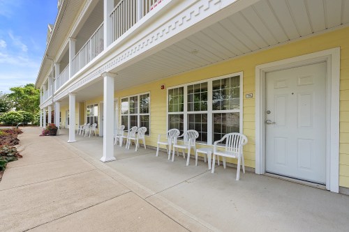 A yellow building with a porch featuring white chairs and tables, surrounded by greenery and a clear blue sky.