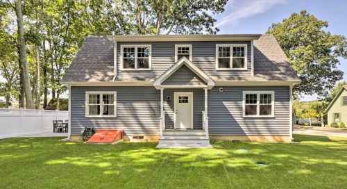 A two-story blue house with a front porch, surrounded by green grass and trees on a sunny day.