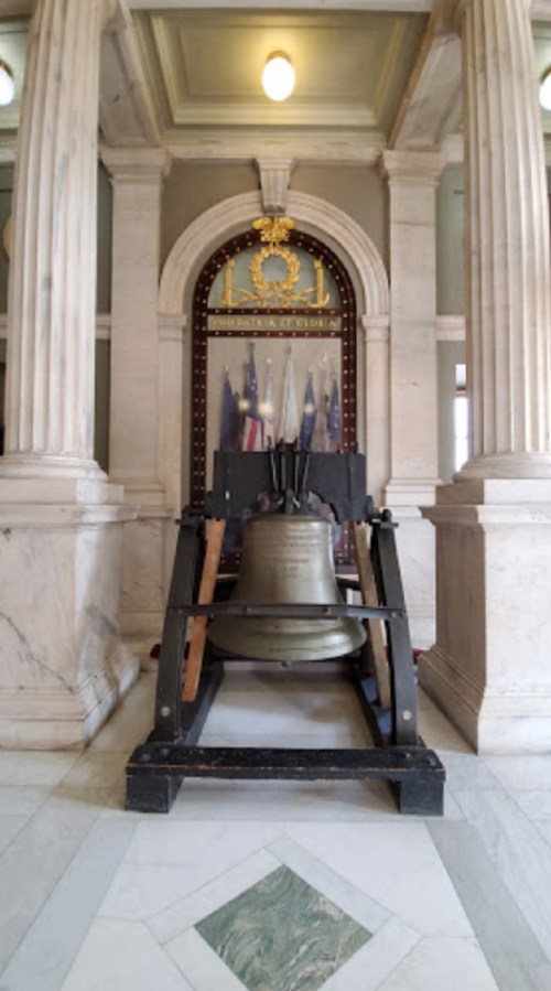 A large bell displayed on a wooden stand inside a marble hall, framed by columns and a decorative backdrop.