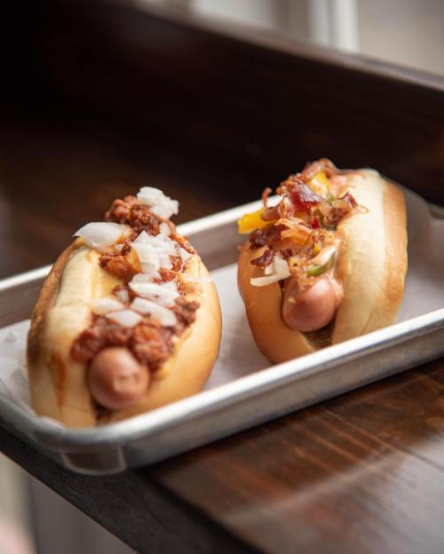 Two hot dogs on a tray, one topped with chili and onions, the other with bacon and diced peppers.
